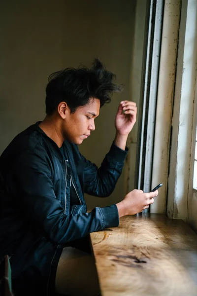 portrait of thoughtful man sitting with smartphone at wooden table near window