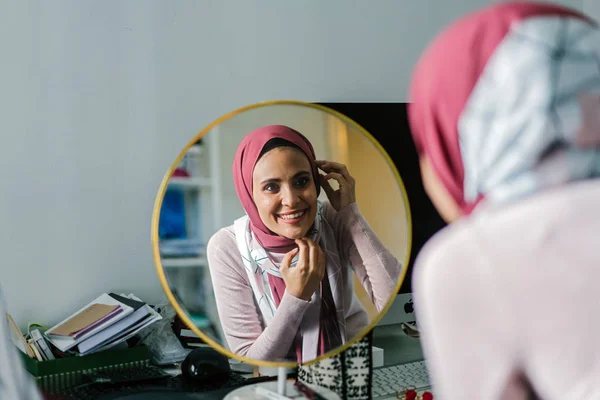 smiling young woman looking at round mirror adjusting her head scarf