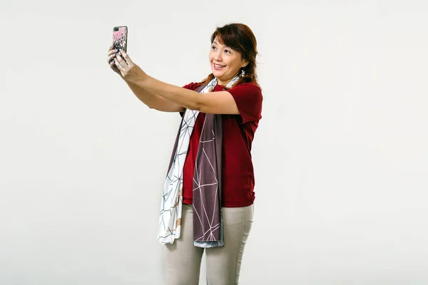asian woman wearing red blouse smiling taking selfie on mobile phone standing on white  studio background