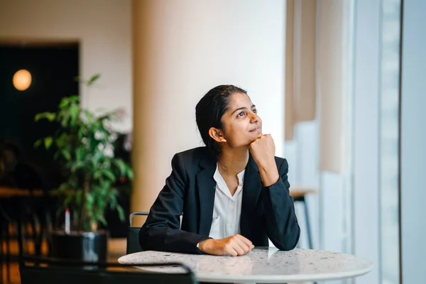 young smiling businesswoman in suit sitting at table and dreaming looking away