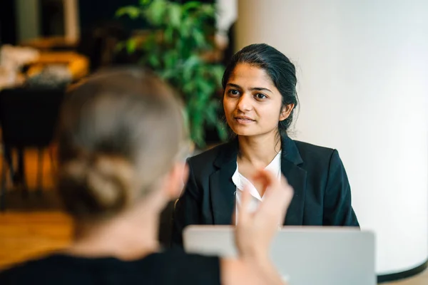 young female business partners smiling and talking sitting at table in modern office