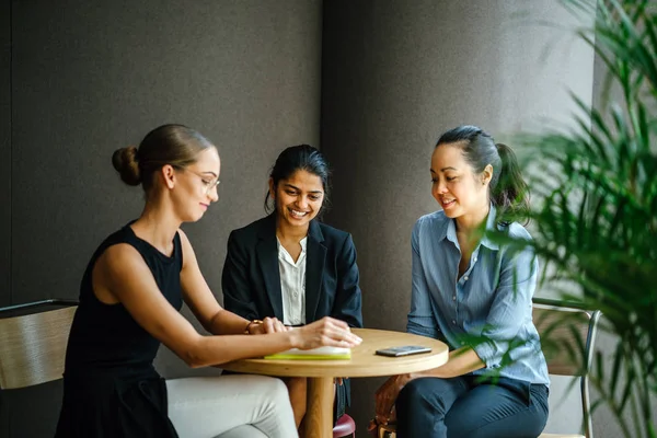 portrait of three young women smiling reading book sitting at table, three quarter length