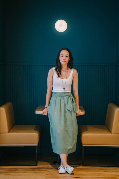 asian woman wearing stylish top, long skirt and sneakers smiling leaning on table 