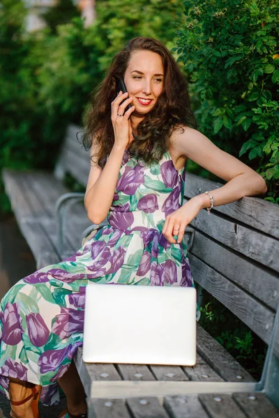 brunette woman with laptop talking on phone sitting on bench in green park