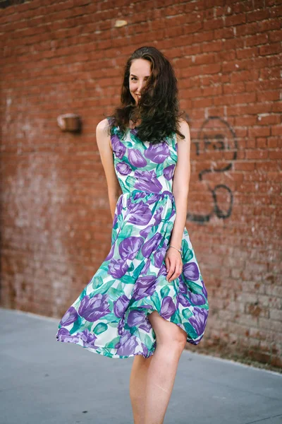 笑みを浮かべて 彼女のドレスは 煉瓦壁の背景でポーズを保持している巻き毛の若くてきれいな女性 — ストック写真