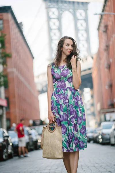 街の通りの上を歩く花印刷保持袋とドレスを着て巻き毛を持つ若い笑顔の女性 — ストック写真