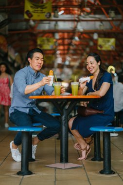 Portrait of a good looking Chinese Asian couple enjoying a snack at a hawker center during the weekend in Singapore, Asia. They are enjoying  sugarcane juice and fried bananas. clipart