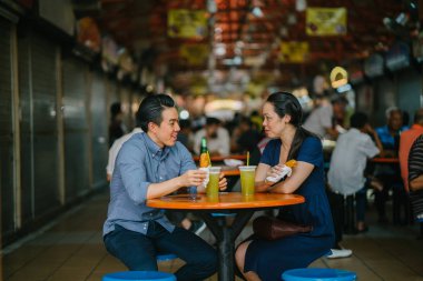 Portrait of a good looking Chinese Asian couple enjoying a snack at a hawker center during the weekend in Singapore, Asia. They are enjoying  sugarcane juice and fried bananas. clipart