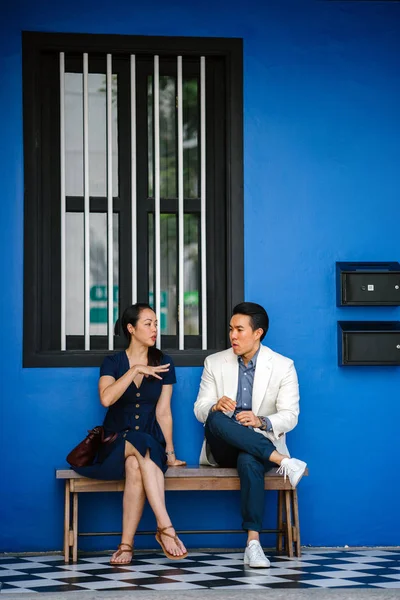 A Chinese Asian couple are out on a date. They are both middle aged, well-dressed, attractive and enjoying a conversation with one another while sitting on a bench at a colorful shop house.