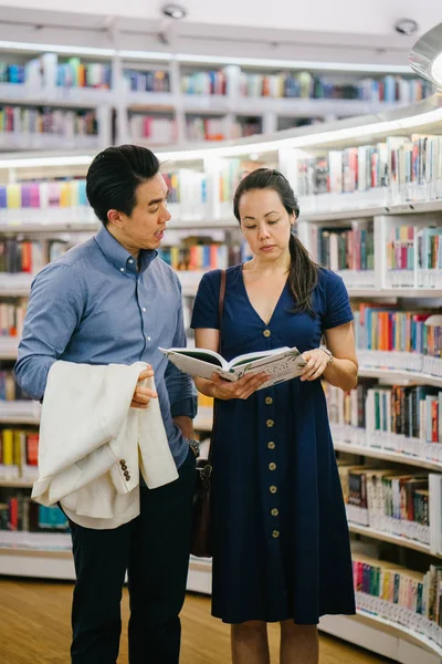 A Chinese Asian couple are on a date. They are reading a book in a library over the weekend. They are smiling as they stand close to one another.