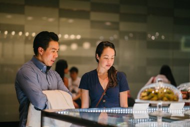  Genç Çinli Asyalı Çift kek bir perakende alışveriş merkezinde bir görüntüsünü düşünüyor. Onlar bir hafta sonu bir tarihte tatlılar sahip düşünüyor musun.