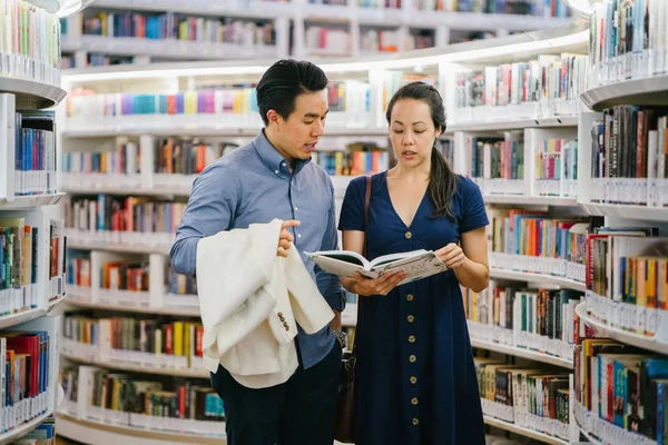 A Chinese Asian couple are on a date. They are reading a book in a library over the weekend. They are smiling as they stand close to one another.