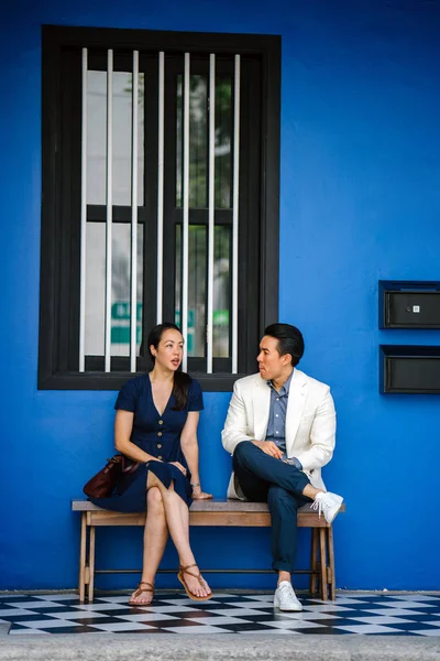 A Chinese Asian couple are out on a date. They are both middle aged, well-dressed, attractive and enjoying a conversation with one another while sitting on a bench at a colorful shop house.