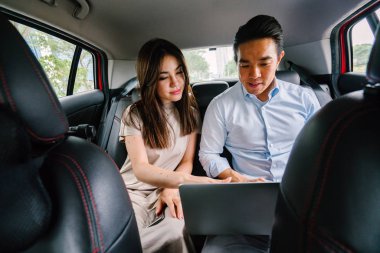 Two young Asian business people sit on the back seat of a car and are driven to their destination on a ride they booked via a ride hailing app. clipart