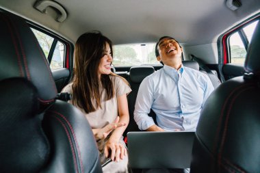 Two young Asian business people sit on the back seat of a car and are driven to their destination on a ride they booked via a ride hailing app. clipart