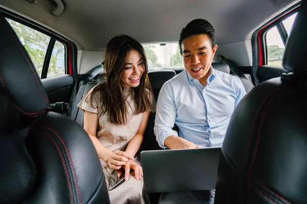 Two young Asian business people sit on the back seat of a car and are driven to their destination on a ride they booked via a ride hailing app.