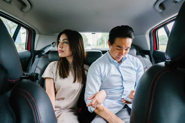 Two young Asian business people sit on the back seat of a car and are driven to their destination on a ride they booked via a ride hailing app.