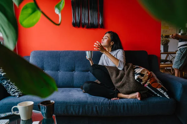 Portrait of a Singaporean Malay woman sitting on a comfortable blue couch with tea in a trendy living room during the daytime. She is relaxing and chilling out on her own on a weekend.