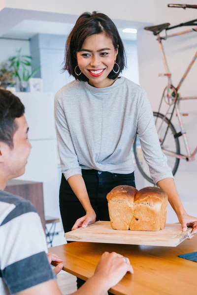 A young Malay woman bakes bread for her husband and serves the freshly baked bread to him on a wooden tray on the weekend