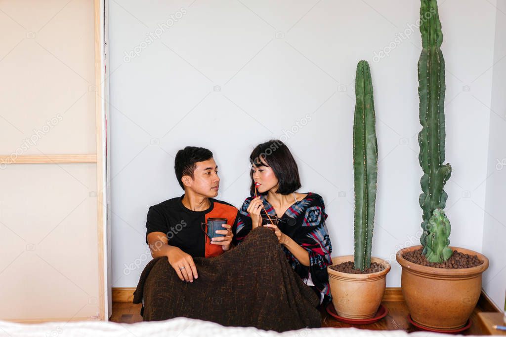 A young Malay couple snuggle up and share a hot beverage in their home. They are wrapped up and sitting on the floor and cosy as they talk to one another.