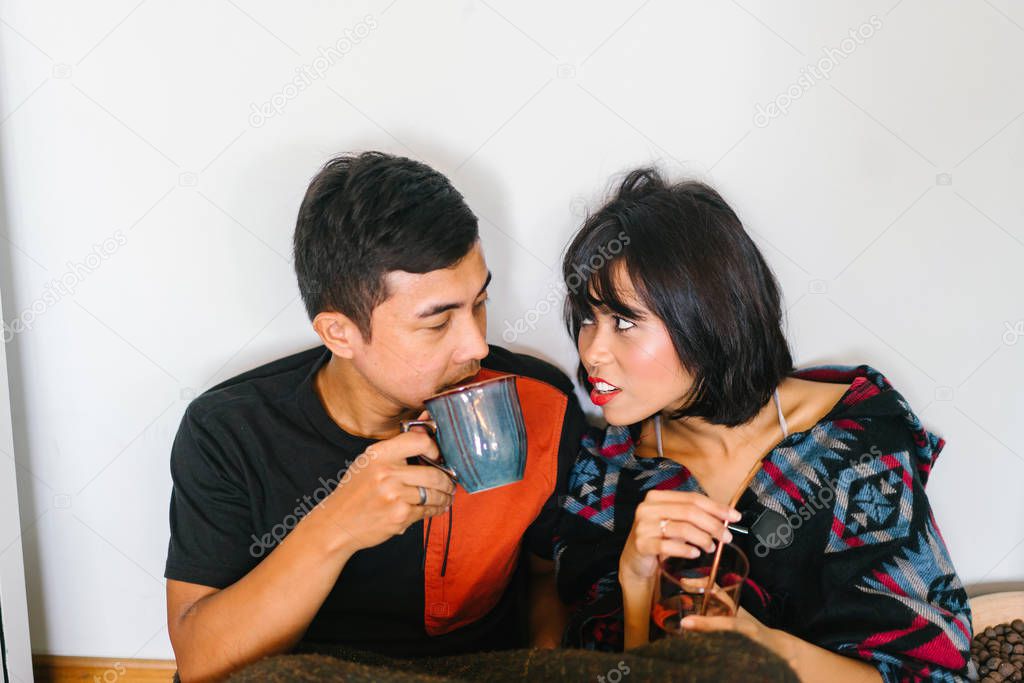 A young Malay couple snuggle up and share a hot beverage in their home. They are wrapped up and sitting on the floor and cosy as they talk to one another.