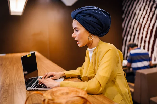 Portrait of a young Muslim Malay woman working or studying on her laptop computer indoors. She is dressed in a modest by stylish pastel outfit and a turban (hijab, head scarf).