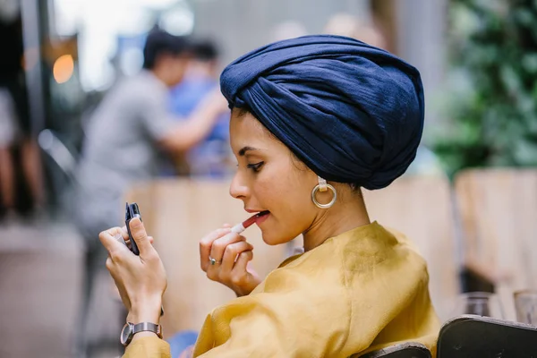 A stylish and young Muslim Malay woman wearing a turban (hijab, head scarf) applying lipstick and makeup in a cafe during the daytime. She is attractive, elegant and fashionably dressed.