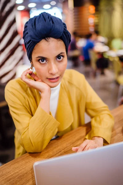 Portrait of a young Muslim Malay woman working or studying on her laptop computer indoors. She is dressed in a modest by stylish pastel outfit and a turban (hijab, head scarf).