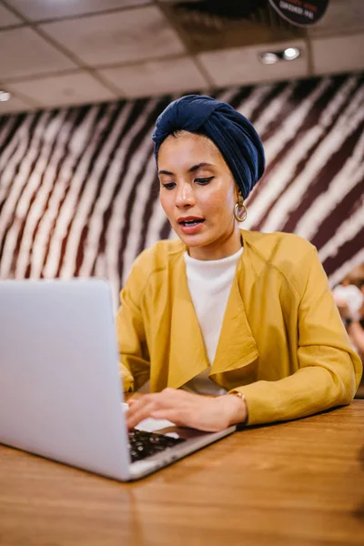 Portrait of a young Muslim Malay woman working or studying on her laptop computer indoors. She is dressed in a modest by stylish pastel outfit and a turban (hijab, head scarf).