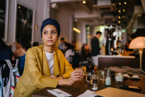 young and elegant Muslim Malay woman with  her coffee in a cafe. She is wearing a stylish outfit and turban (hijab, head scarf) and earrings.