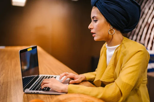 Portrait of a young Muslim Malay woman working or studying on her laptop computer indoors. She is dressed in a modest by stylish pastel outfit and a turban (hijab, head scarf).