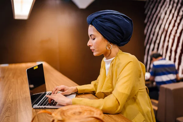 Portrait of a young Muslim Malay woman working or studying on her laptop computer indoors. She is dressed in a modest by stylish pastel outfit and a turban (hijab, head scarf).