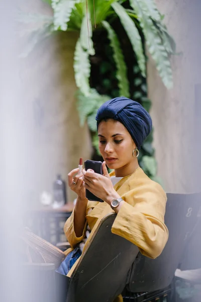 A stylish and young Muslim Malay woman wearing a turban (hijab, head scarf) applying lipstick and makeup in a cafe during the daytime. She is attractive, elegant and fashionably dressed.