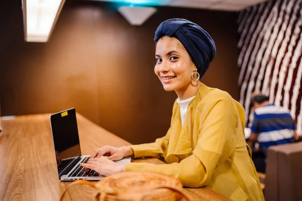 Portrait of a young Muslim Malay woman working or studying on her laptop computer indoors. She is dressed in a modest by stylish pastel outfit and a turban (hijab, head scarf).