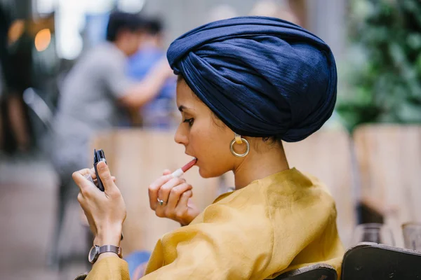 A stylish and young Muslim Malay woman wearing a turban (hijab, head scarf) applying lipstick and makeup in a cafe during the daytime. She is attractive, elegant and fashionably dressed.