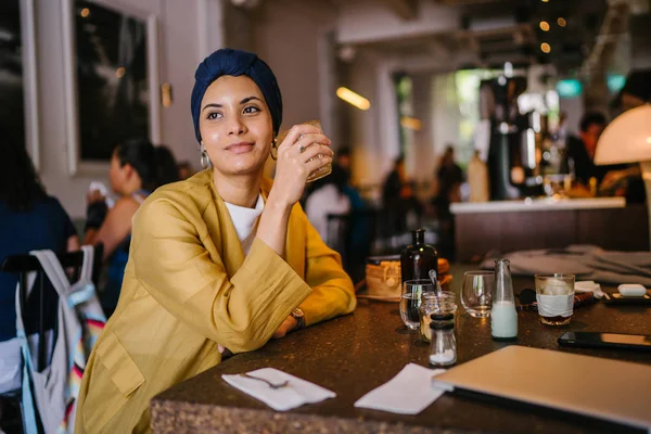 young and elegant Muslim Malay woman with  her coffee in a cafe. She is wearing a stylish outfit and turban (hijab, head scarf) and earrings.