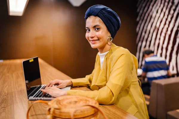 Portrait of a young Muslim Malay woman working or studying on her laptop computer indoors. She is dressed in a modest by stylish pastel outfit and a turban (hijab, head scarf).