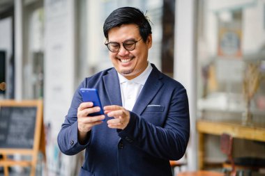 Orta yaşlı Çinli Asyalı portresi adam sıradan bir takım elbise (ceket, oxford düğme aşağı gömlek ve kot) ve onun smartphone kullanarak gözlük