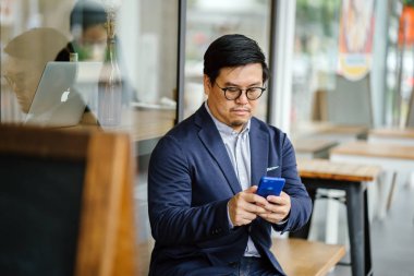 Orta yaşlı bir Çinli Asyalı adam rahat takım elbise (ceket, oxford düğme aşağı gömlek ve kot) ve gözlük bir kafede şehir dışında bir bankta oturur gibi onun smartphone kullanarak portresi.