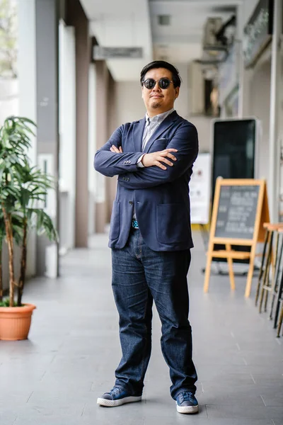 Portrait of a middle-aged Chinese Asian man (Singaporean) in smart casual standing in the city. He is well-dressed and groomed in a navy suit, oxford button-down shirt  with sunglasses.