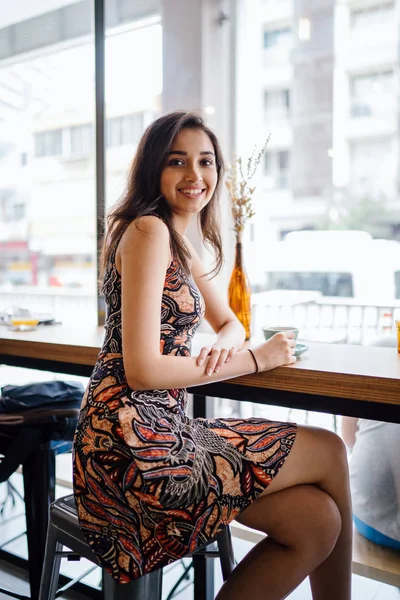 Young and beautiful Indian Asian Singaporean woman in a print dress sits in a cafe as she enjoys the weekend in Asia.