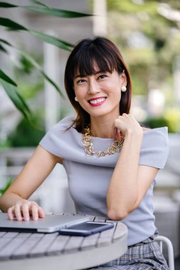 Açık havada şık bir yerde oturan bir emin, şık ve çekici orta yaşlı Çinli Asyalı kadın portresi. O bir pohpohlama gri kıyafeti giyiyor.