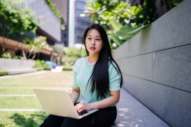 A young and attractive Asian woman student sits and works on her laptop computer on a university campus during the day. She is casually dressed as she studies.  clipart