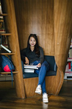 Genç, güzel ve çekici Çinli Asyalı değişim öğrencisi kütüphanedeki kahverengi bir kulübede okuyor ve çalışıyor..