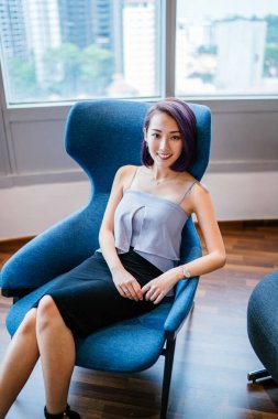 Profesyonel elbiseli ve boyalı mor saçlı genç, ince ve zarif bir Asyalı kadın çalışma odasında kırmızı bir koltukta oturuyor. İçtenlikle gülümsüyor..