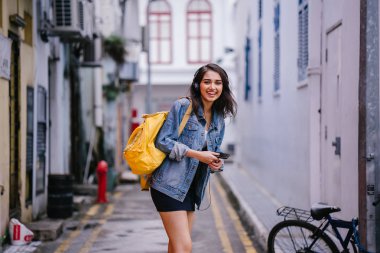 Bir genç ve çekici Hint Asyalı kadın bir kot ceket ve sarı sırt çantası onun kulaklıklar üzerinde müzik keyfi portresi