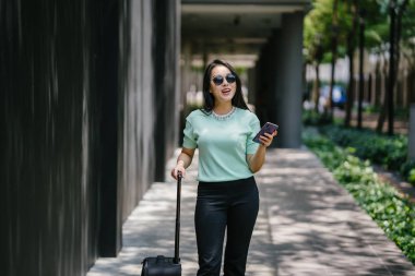 O bir yolculuk dolu uygulama üzerinden rezervasyonu onu binmek için caddenin kenarında bekleyen zarif ve güzel bir genç Asyalı kadının portresi. Tekerlekli bir bavula tutunuyor.. 