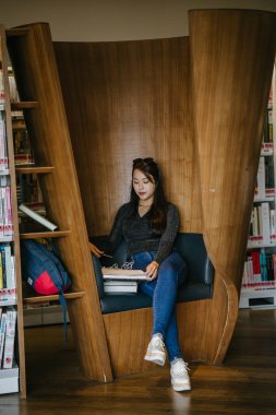 Genç, güzel ve çekici Çinli Asyalı değişim öğrencisi kütüphanedeki kahverengi bir kulübede okuyor ve çalışıyor..