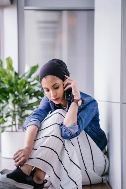 Genç bir Ortadoğulu Müslüman kadın gün boyunca bir ofiste, ortak çalışma alanında veya kütüphanede oturuyor ve akıllı telefonunu kullanıyor. Başörtüsü takıyor (başörtüsü)).
