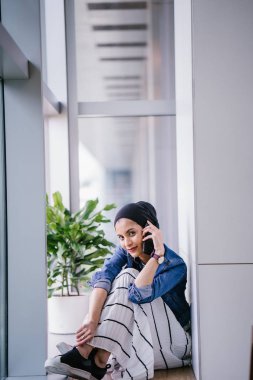 Genç bir Ortadoğulu Müslüman kadın gün boyunca bir ofiste, ortak çalışma alanında veya kütüphanede oturuyor ve akıllı telefonunu kullanıyor. Başörtüsü takıyor (başörtüsü)).